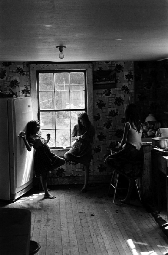 Three girls in a dimly lit kitchen, one leaning on a fridge, another by a window, and the third sitting on a counter, all in casual poses.