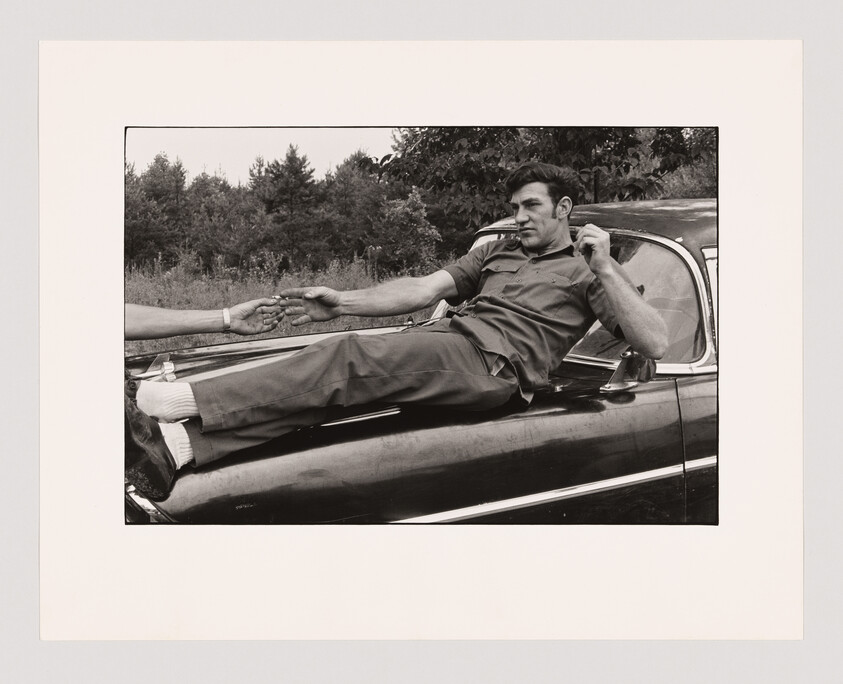 Man in work clothes lies on a car hood, reaching out to touch another person’s hand. Trees in background.