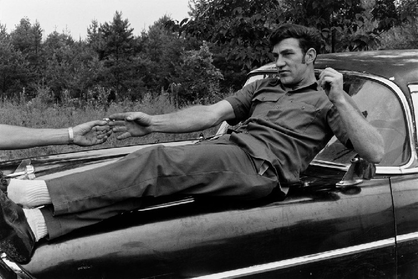 Man reclining on a car hood, reaching out to another person. Trees in the background. Casual outdoor setting.