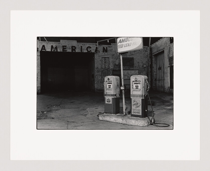 Two old gas pumps stand in front of a worn building with a faded sign reading "AMERICAN."