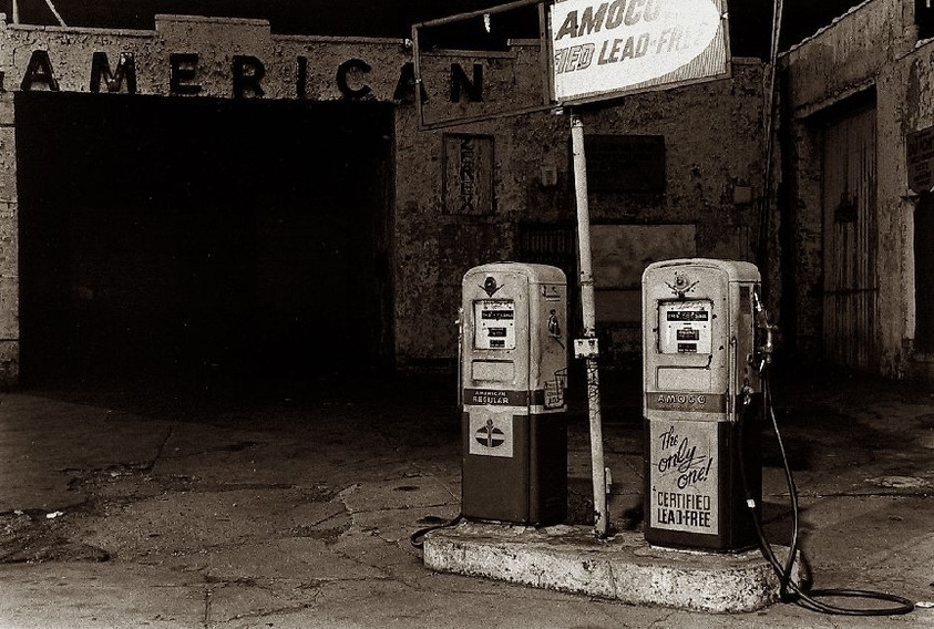 Vintage gas station with two old pumps, one labeled "Amoco" and "Certified Lead-Free." The word "American" is partially visible in the background.