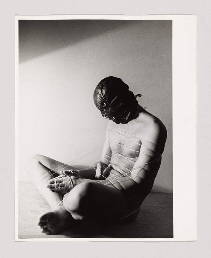 A person sits on the floor, nude, with their head covered and body wrapped in string or cord.