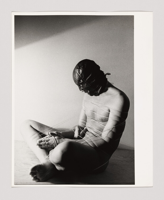 Naked person wearing a leather hood sits bound by string, hands tied and head bowed.