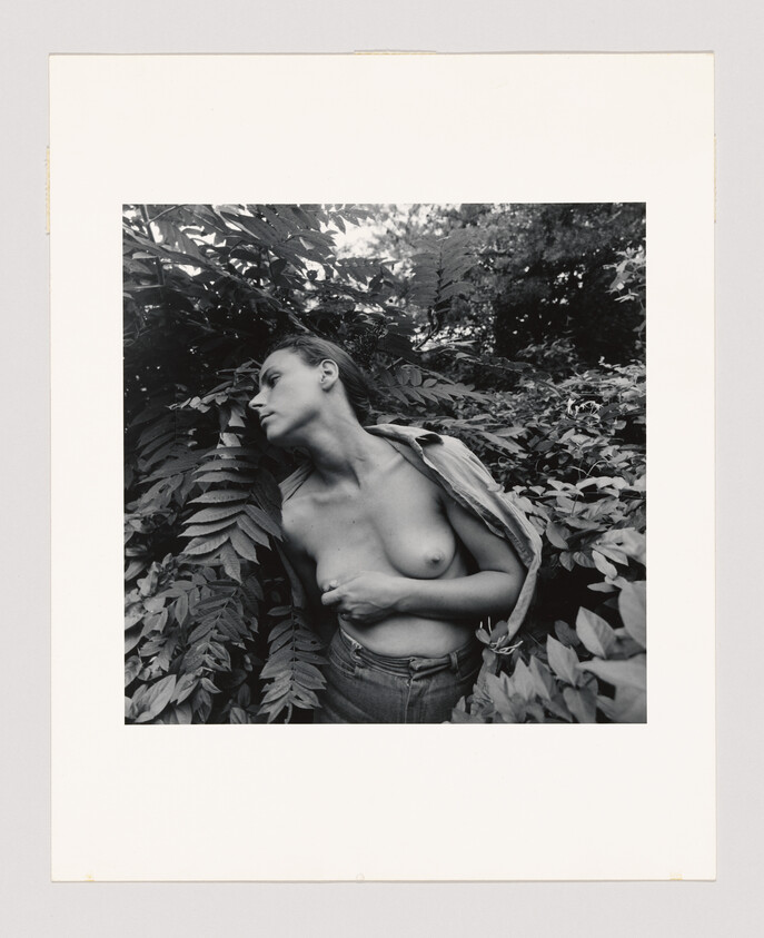 A topless person stands among dense leafy plants, looking to the side with one arm across their chest.