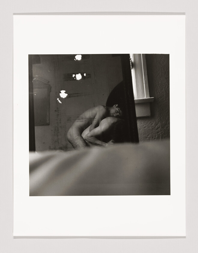 Two nude people embrace closely in a dimly lit room, partially reflected in a mirror.