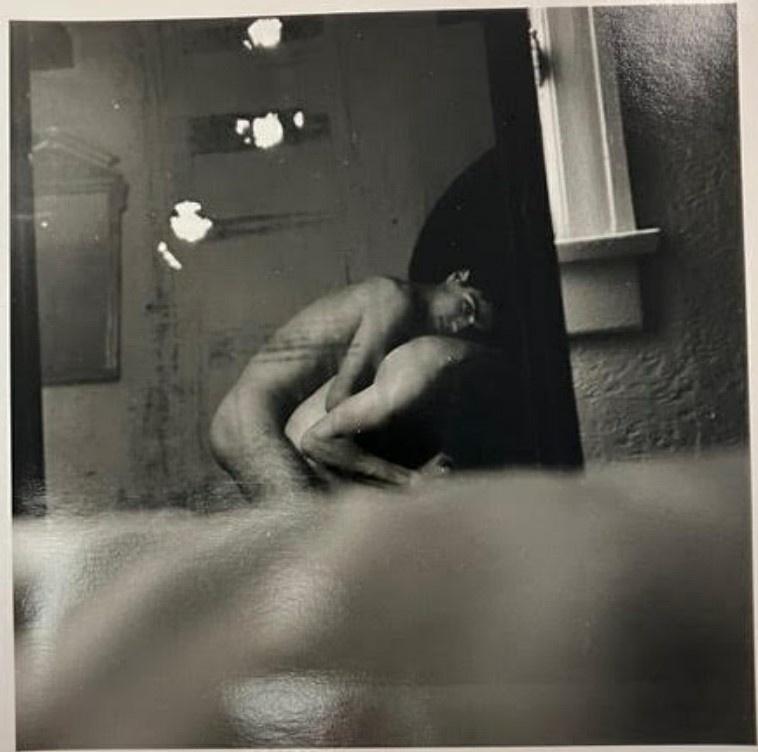 Two nude individuals are embracing in a dimly lit room with a window and a mirror on the wall. The image is in black and white.