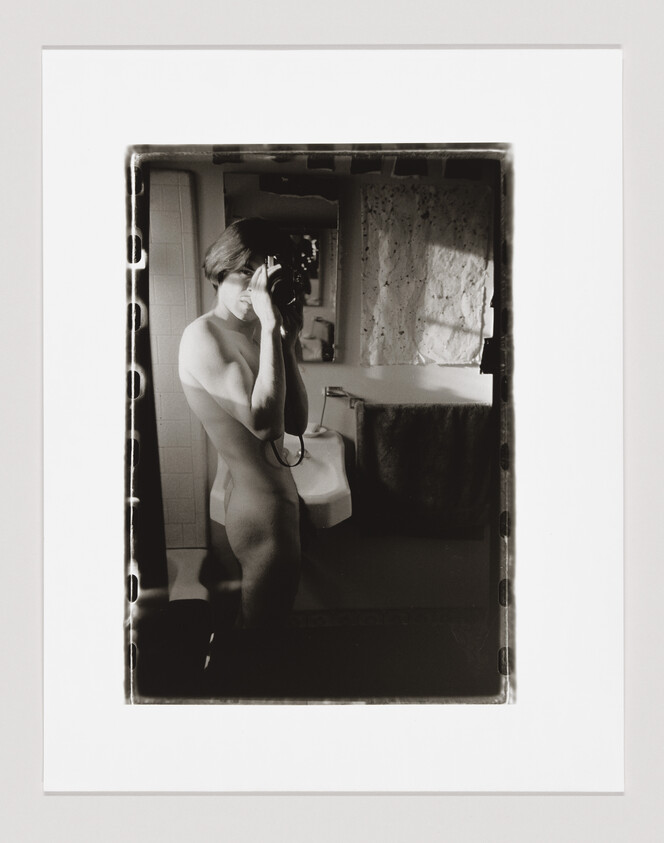 A nude person stands in a bathroom, taking a self-portrait with a camera in front of a mirror.