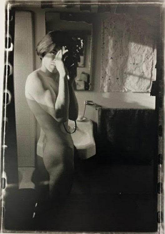 A black-and-white photo of a person taking a mirror selfie in a bathroom, holding a camera, and standing nude.