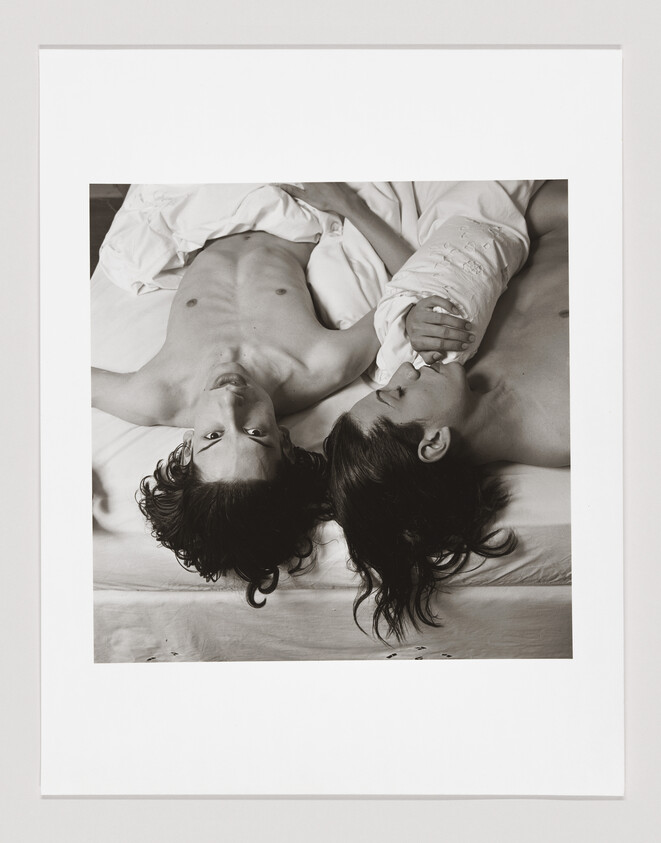 Two people with long hair lie side by side in bed, holding hands and looking in different directions.