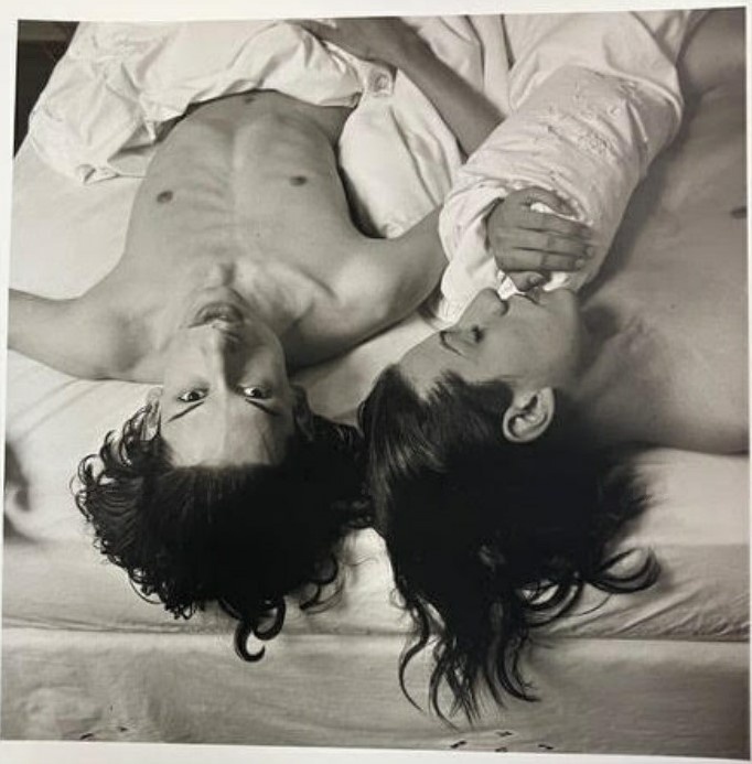 Two people with long hair lie on a bed, facing opposite directions. One looks at the camera while the other gazes at the first person.