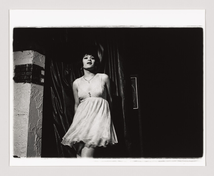 Person in a white dress stands on stage, partially illuminated, with a curtain in the background.