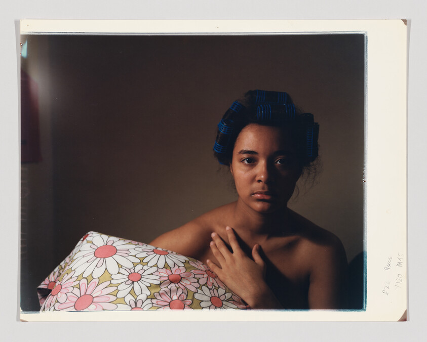 Young woman with blue hair rollers holds a floral pillow and looks directly ahead.