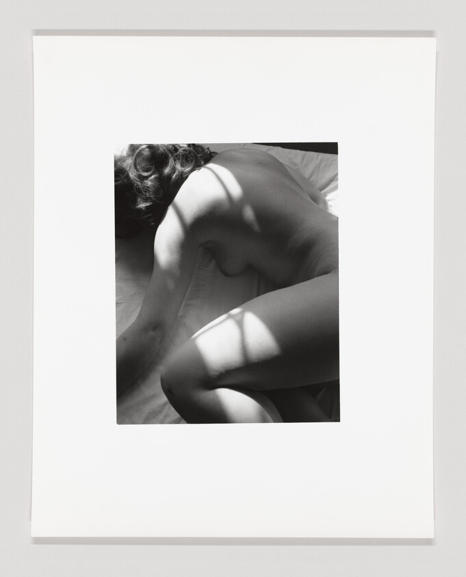 Nude person curled on a bed with window shadows falling across their back and legs.