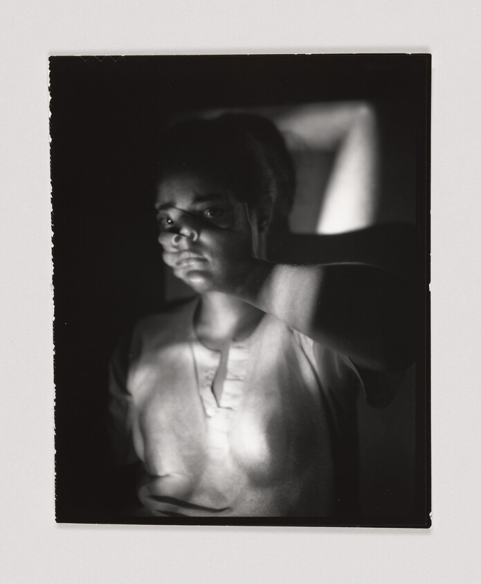 A person stands in dim light with their hand covering part of their face, looking directly forward.