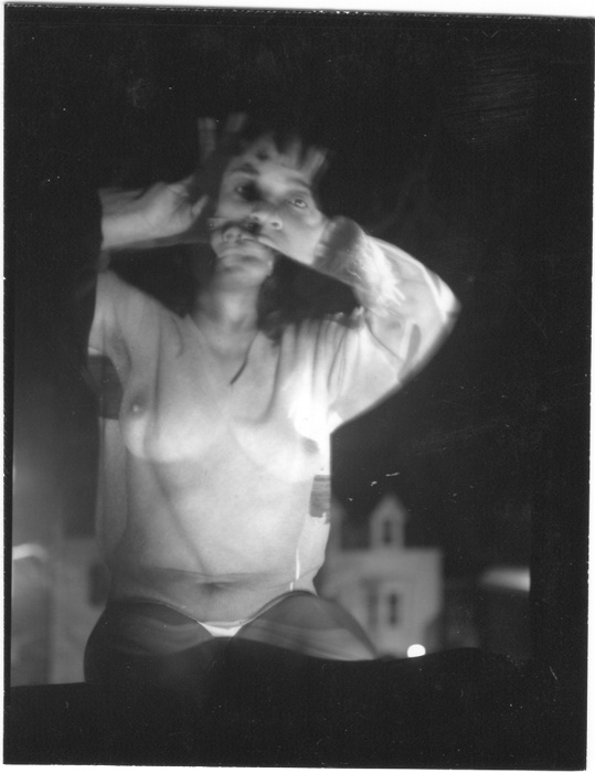 A woman in a white top and underwear appears in a double exposure effect, with her hands near her face, against a dark background with faint building outlines.