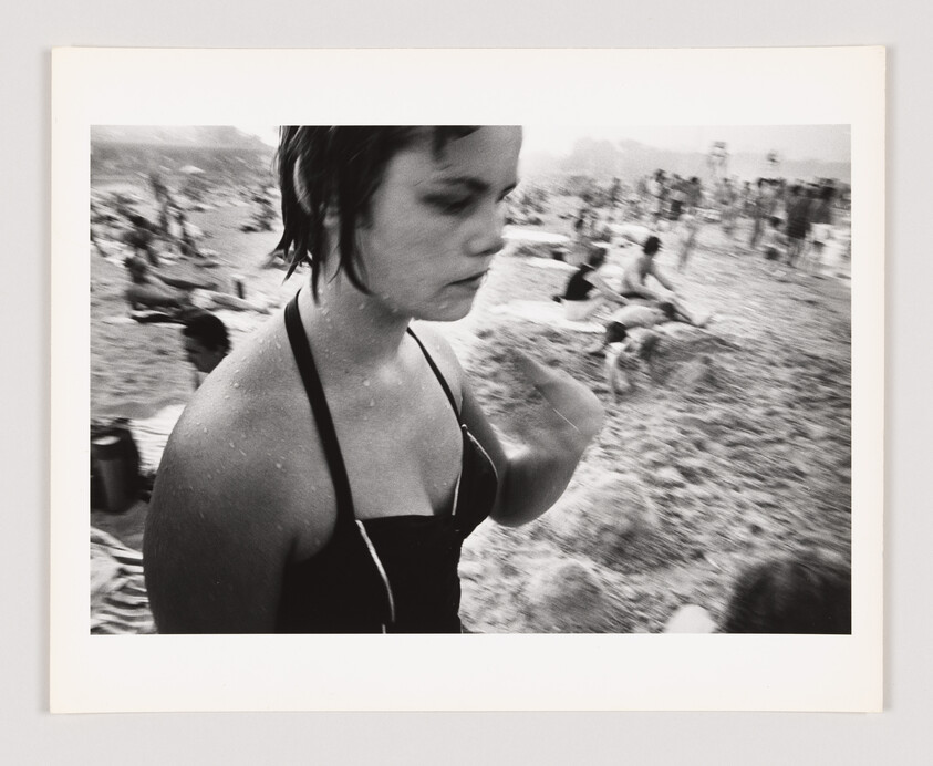 A young woman in a swimsuit stands on a crowded beach, with people sitting and lying on the sand.