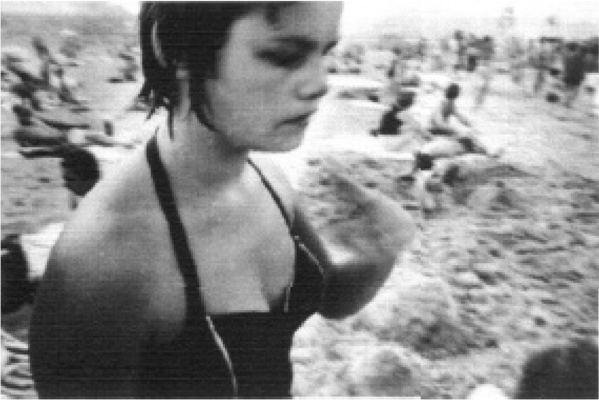 A woman in a swimsuit stands on a crowded beach, looking down. The image is black and white with a grainy texture.