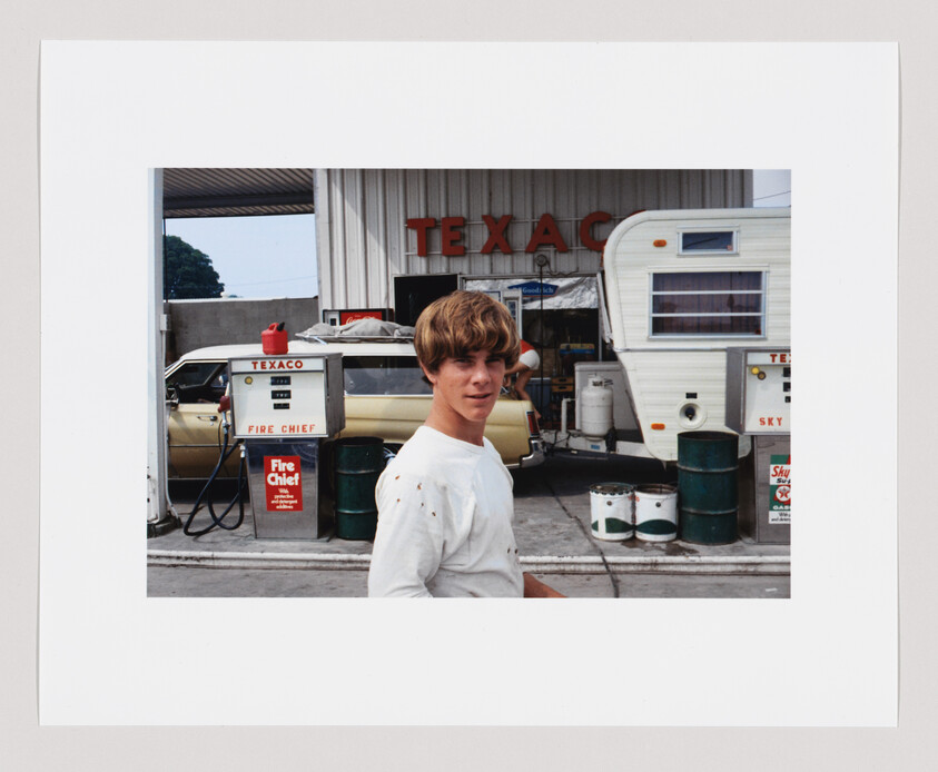 A teenage boy stands in front of a vintage Texaco gas station beside a car and camper.