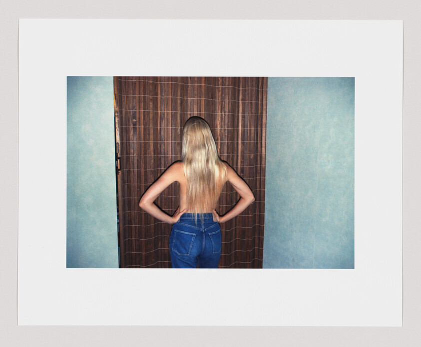 A topless woman with long blonde hair stands with hands on hips facing a wooden curtain.