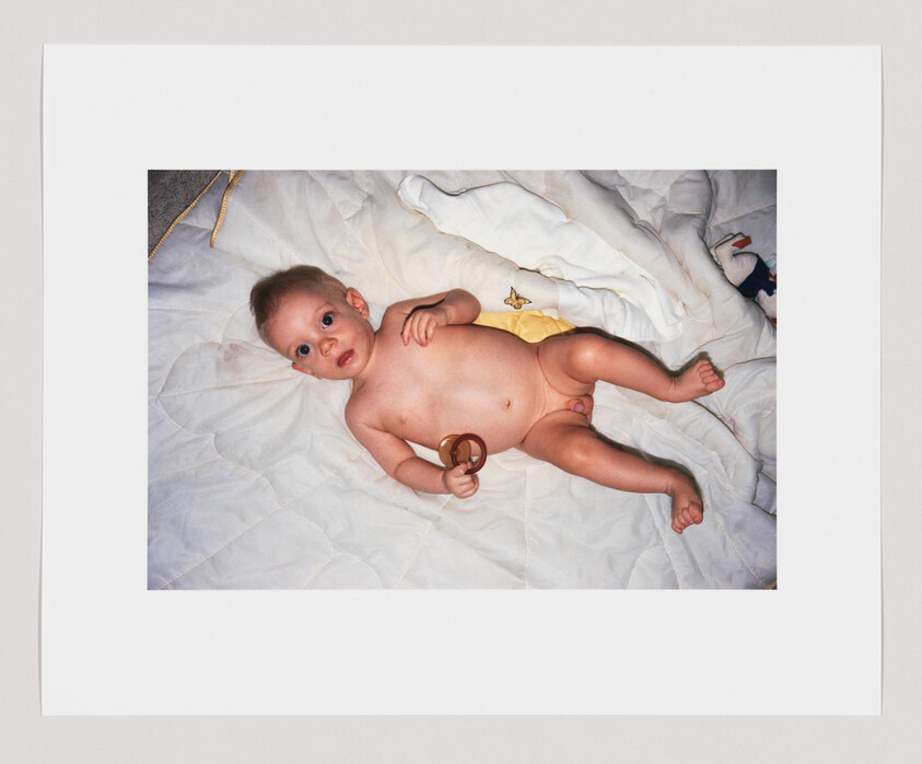 A naked baby lies on a white quilt holding a round wooden rattle and looking up.