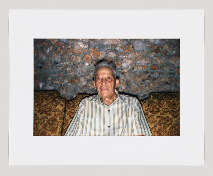 An older man with gray hair sits on a patterned couch, wearing a striped shirt, facing forward.