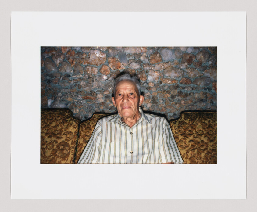 Elderly man sitting on a patterned couch against a stone wall, looking directly at the camera.
