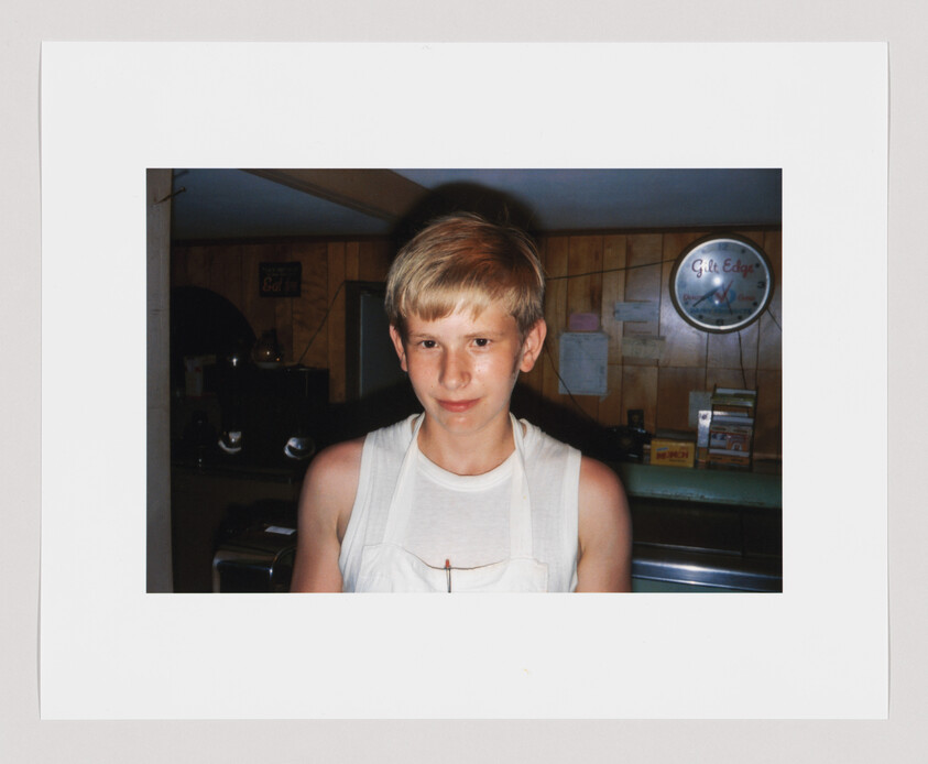 A young person wearing a white tank top stands in a wood-paneled kitchen looking at the camera.
