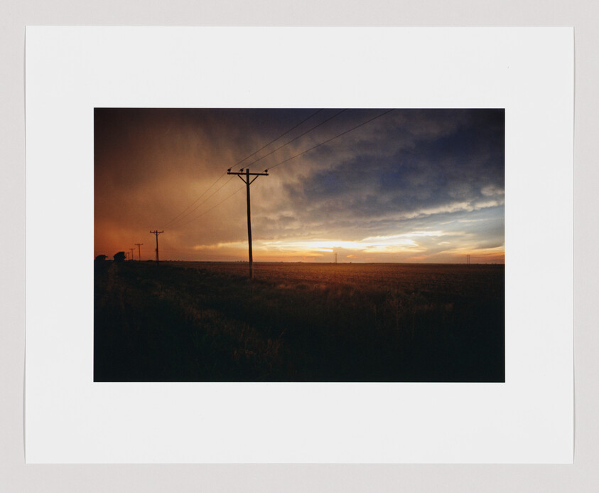 A line of telephone poles crosses a wide field under dramatic sunset clouds.