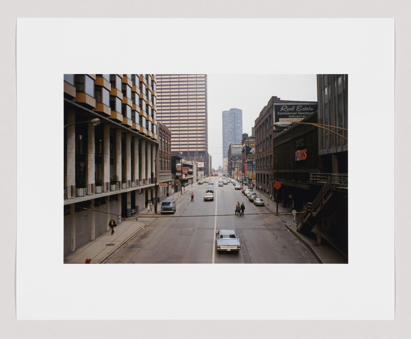 Downtown street with cars driving between tall office buildings and a few pedestrians crossing.