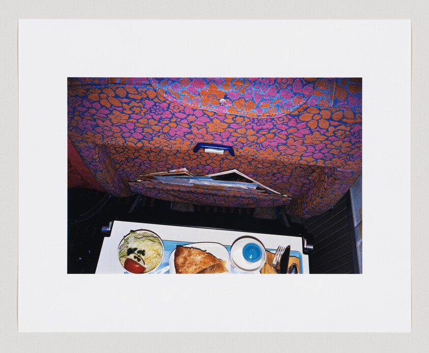 A meal tray with salad, pastry, and cup on a folded tray below a patterned seat.