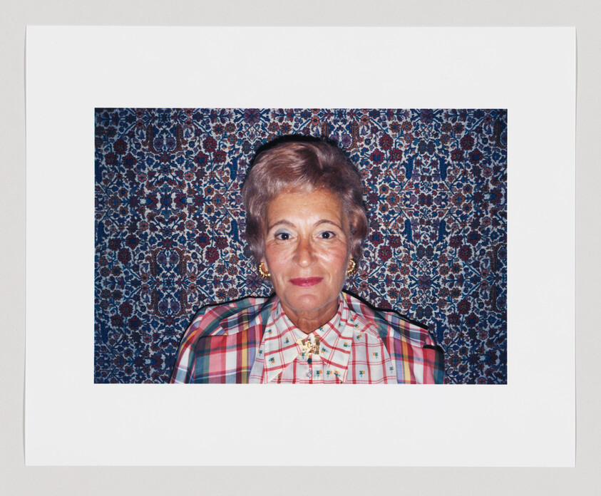 An older woman with short gray hair and patterned blouse posed against a floral tapestry background.