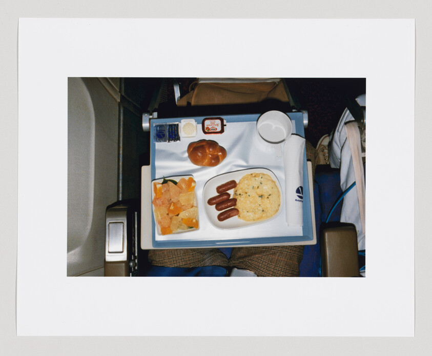 Airplane tray with breakfast: omelette, three small sausages, fruit cup, roll, and empty cup.