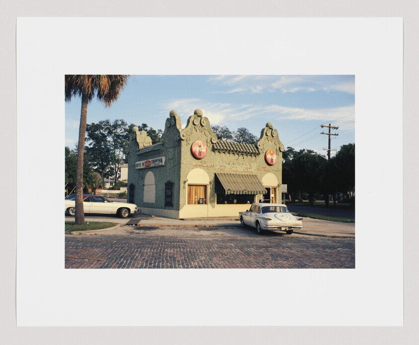 Small vintage diner with Coca-Cola signs, striped awning, and two classic cars parked outside on a brick street.