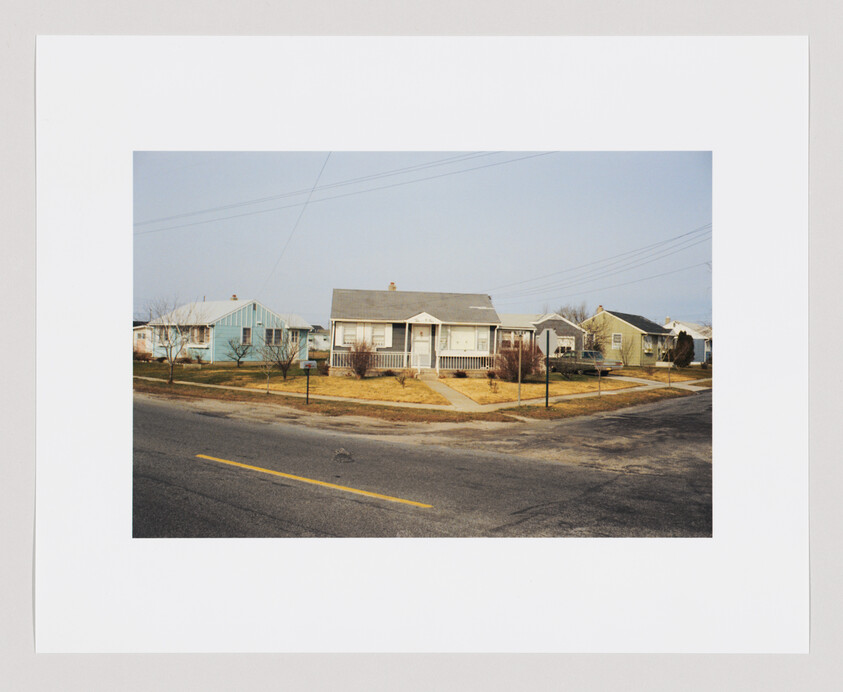 A small house with a front porch sits on a corner lot, surrounded by dry grass and quiet streets.
