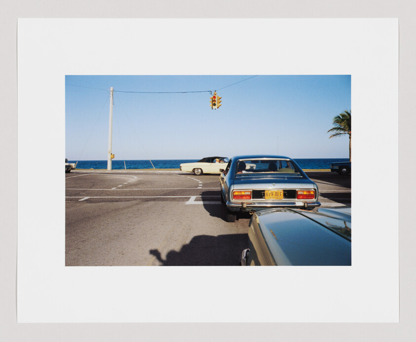 Two vintage cars wait at a seaside intersection under a yellow traffic light with ocean behind.