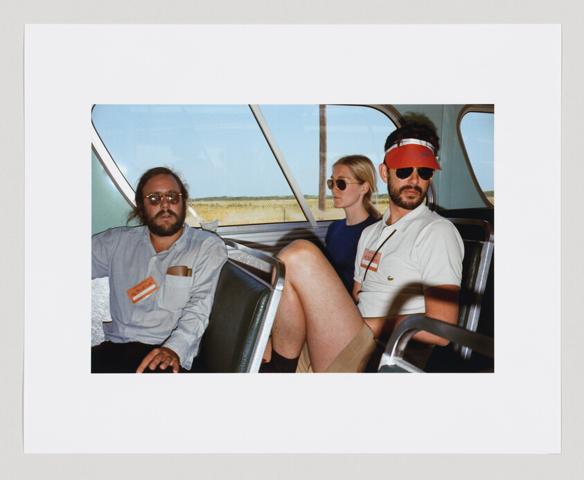 Three people sit on a bus wearing sunglasses, one man in a red visor looking sideways.