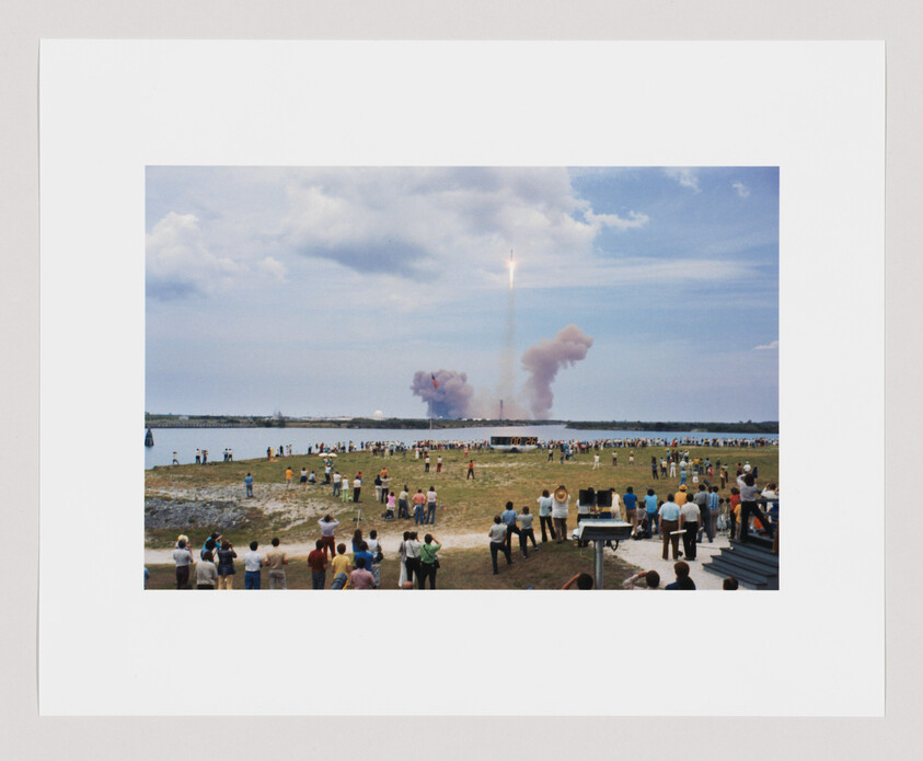 A large crowd watches a rocket launch into the sky, leaving a trail of smoke behind it.