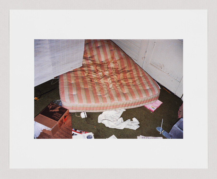 A stained striped mattress on the floor surrounded by scattered clothes, boxes, and papers.