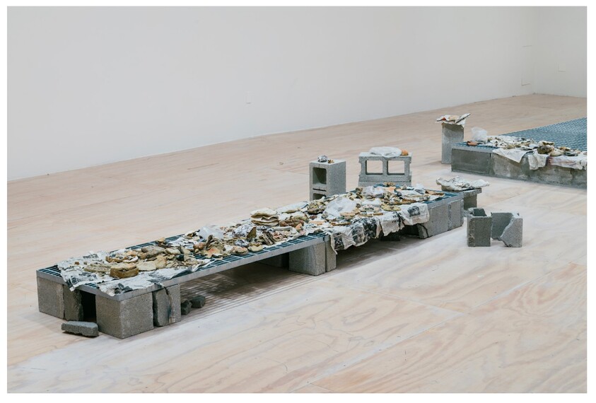 Low sculptural installation of cinder blocks and metal grates covered in torn paper and debris.