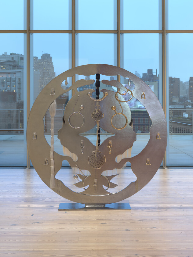 A large round metal sculpture with mirrored face cutouts and hanging rings displayed before floor-to-ceiling windows.