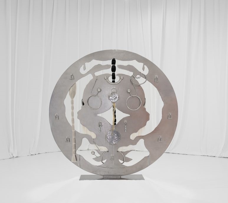 A large, circular metal sculpture with intricate cutouts and attached objects, set against a white draped background.