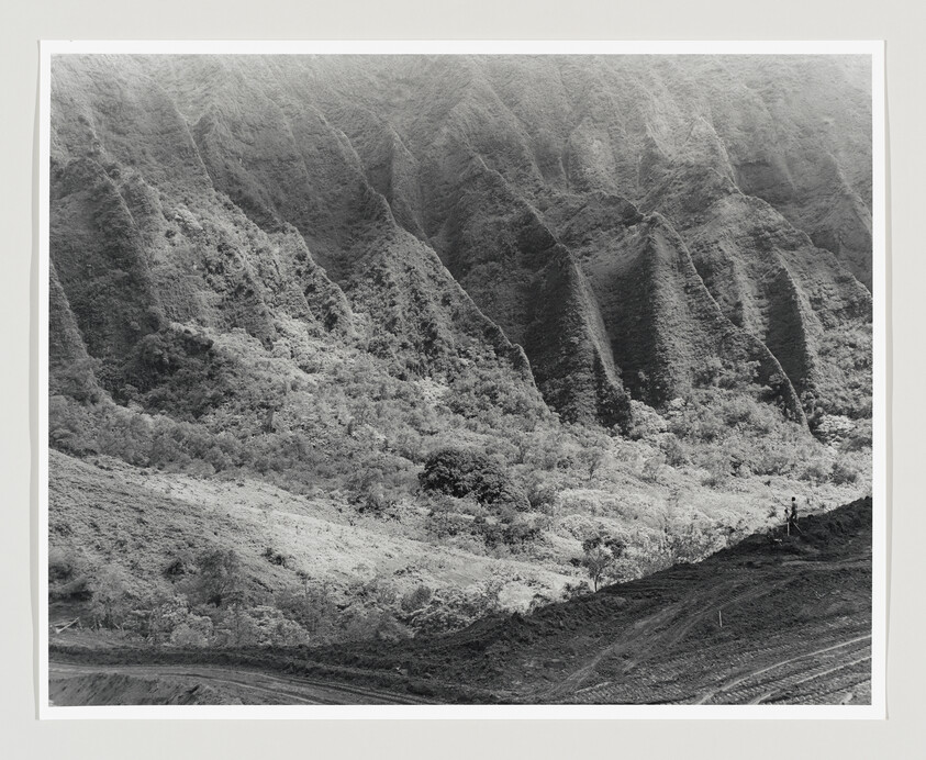 Steep eroded cliffs rise above a vegetated valley with two people walking along a dirt ridge.