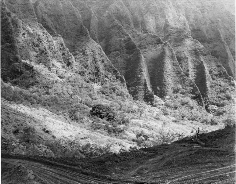 Steep, rugged hills with dense vegetation and a person standing on a dirt path in the foreground.