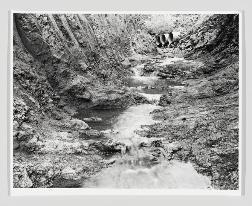 A narrow stream flows through an eroded rocky channel toward three large drainage pipes in the bank.