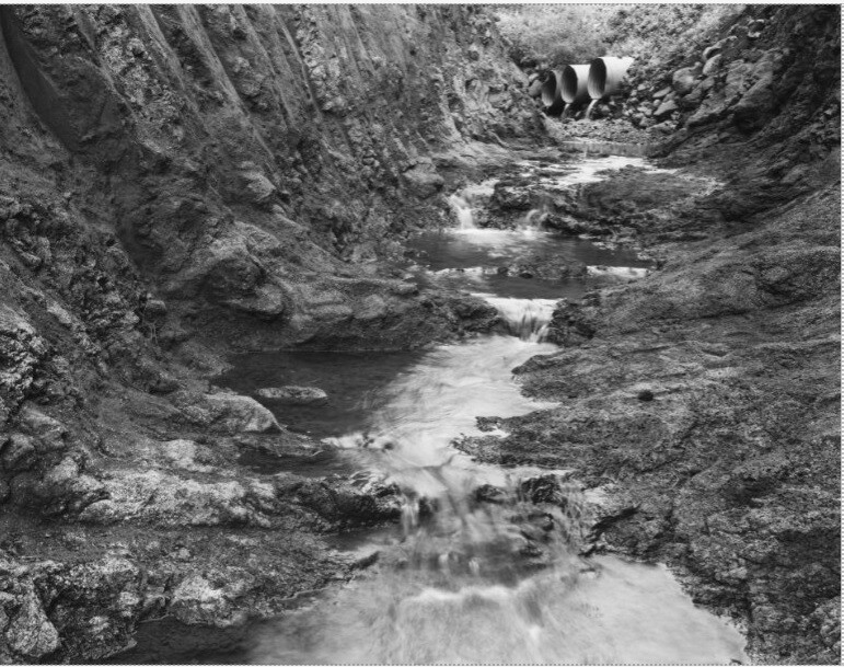 A narrow stream flows through an eroded rocky channel toward three large drainage pipes in the bank.