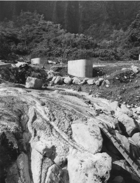Concrete foundations and large rocks in a construction site surrounded by dense greenery and hills.
