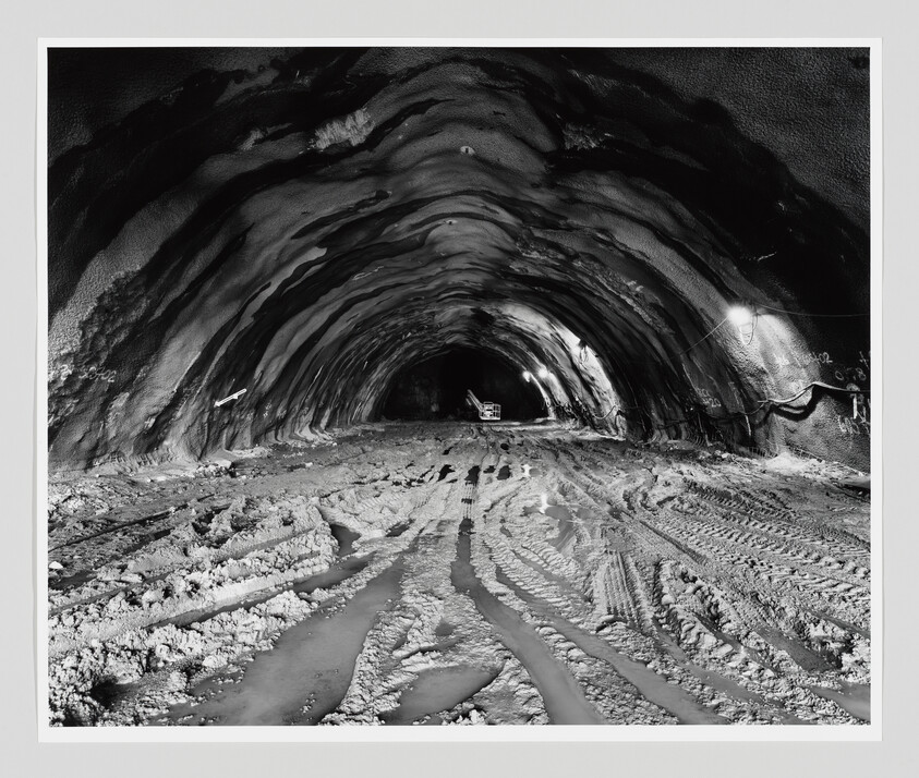 Underground tunnel under construction with muddy vehicle tracks and a small construction vehicle at the far end.