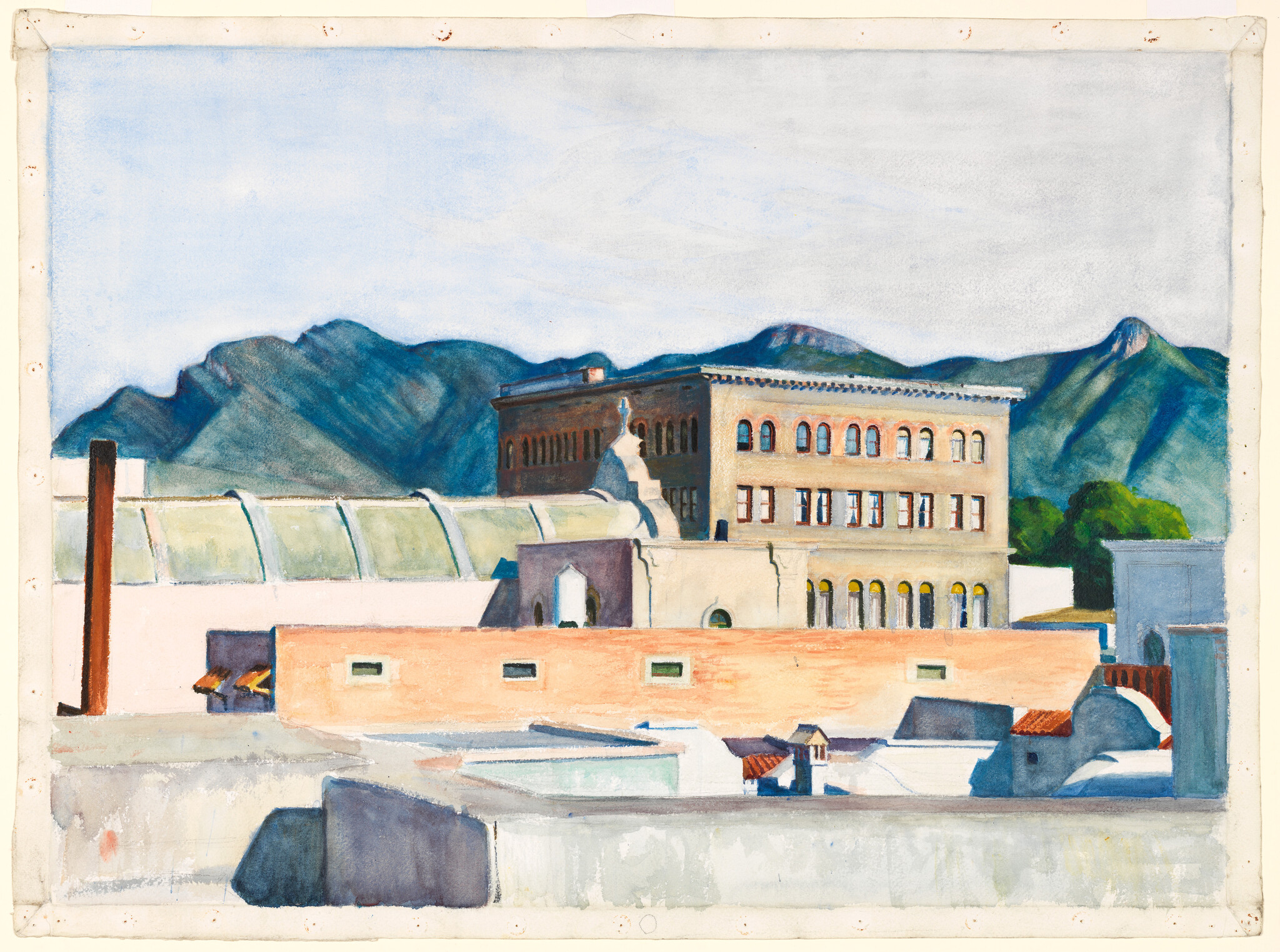 Rooftops and a large brick building sit in front of green mountains under a pale sky.