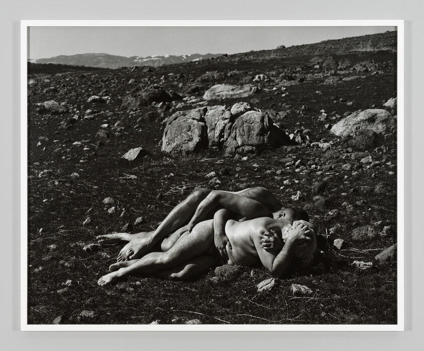 Two naked people lie entwined on rocky ground, holding each other tightly.