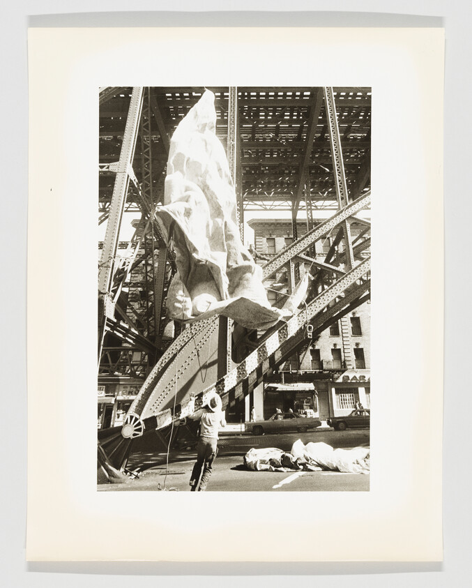 A worker pulls a large billowing fabric under an elevated steel bridge in a city street.
